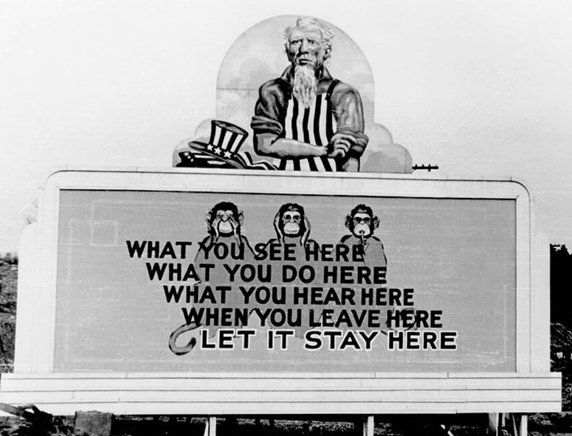 The billboard shows three monkeys and text reading: What you see here, what you do here, what you hear here, when you leave here, let it stay here. On top of the billboard is an old man with gray hair, long gray beard and black-and-white vertically striped shirt and a hat by him side with the same pattern.